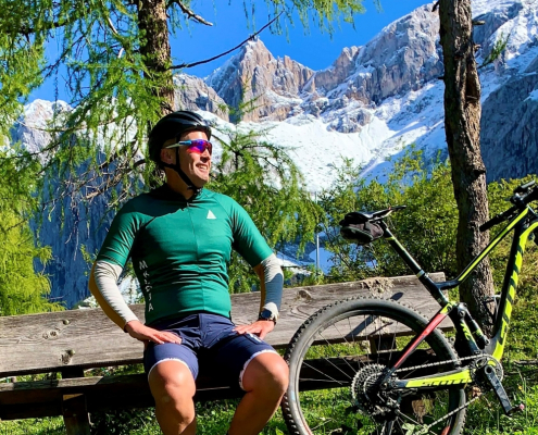 Roland in grünen Radtrikot mit Helm und Brille, sitzt auf einer Bank und lächelt. Neben ihm sein Rad, angelehnt an die Bank. Im Hintergrund Bäume und ein beeindruckender Berg, bedeckt mit Schnee.