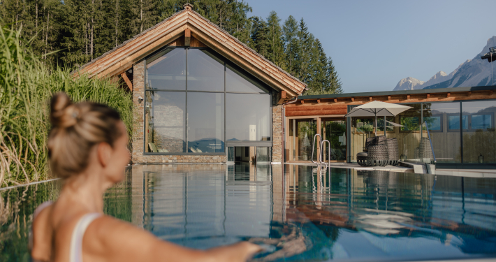 Hotel Annelies Pool Eine Frau blickt von einem Pool aus auf ein modernes Holz- und Steingebäude mit großen Glasfronten, umgeben von Bäumen und Bergen.