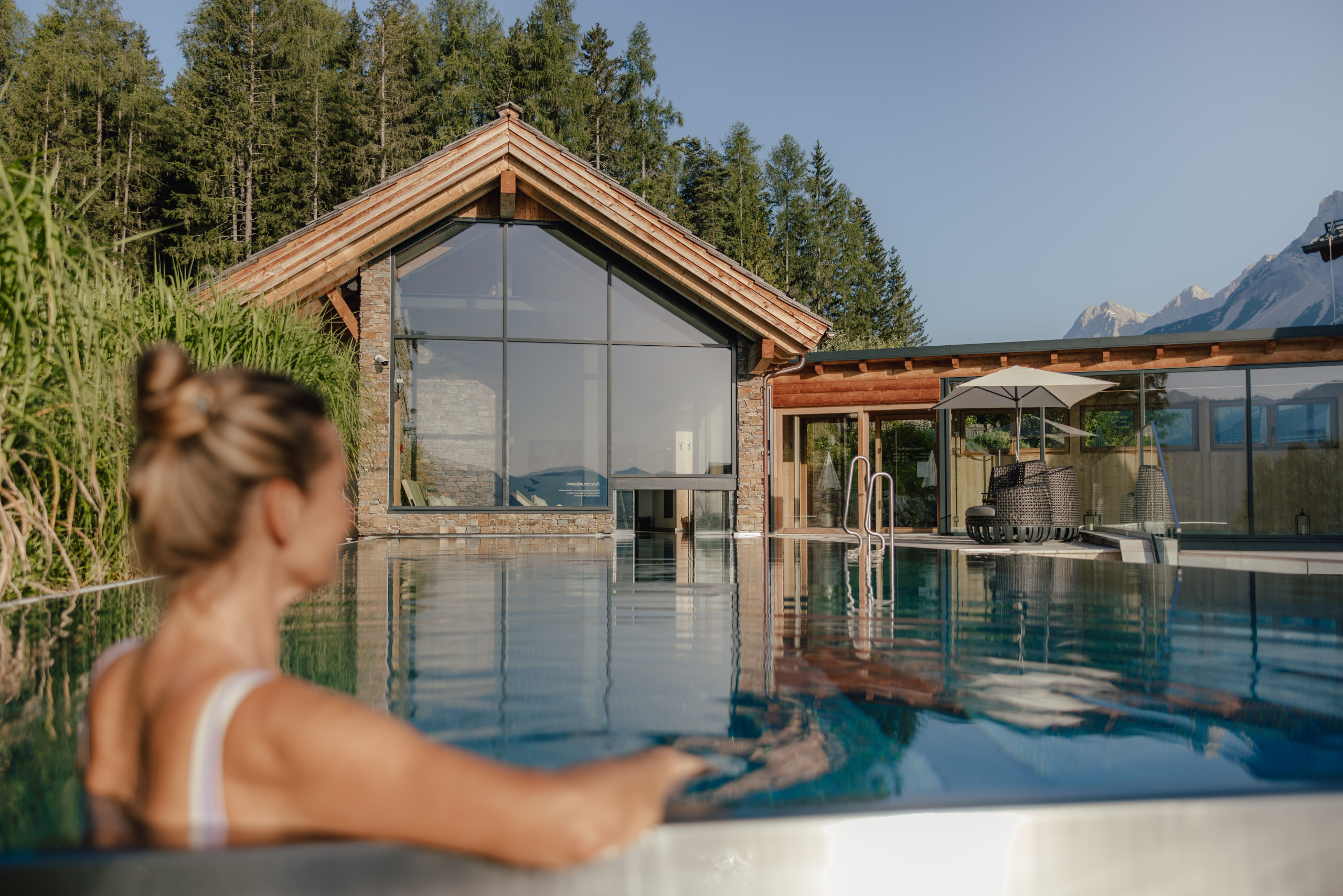 Eine Frau blickt von einem Pool aus auf ein modernes Holz- und Steingebäude mit großen Glasfronten, umgeben von Bäumen und Bergen.