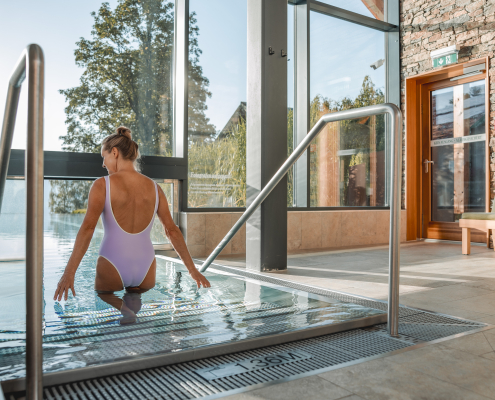 Eine Frau in einem lilafarbenen Badeanzug steigt über eine Treppe in ein Schwimmbecken. Im Hintergrund sind große Fenster und eine Steinwand zu sehen.