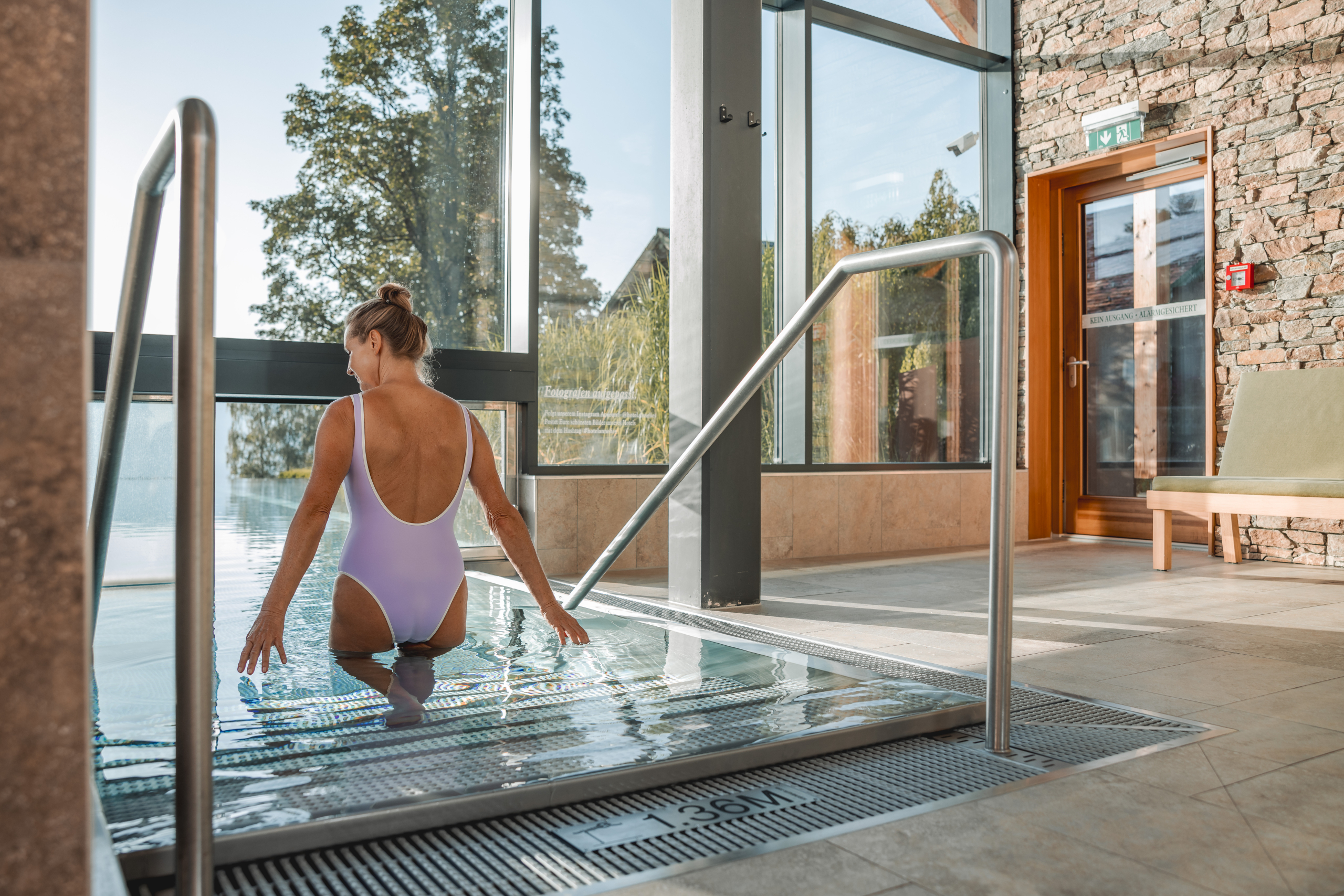 Eine Frau in einem lilafarbenen Badeanzug steigt über eine Treppe in ein Schwimmbecken. Im Hintergrund sind große Fenster und eine Steinwand zu sehen.