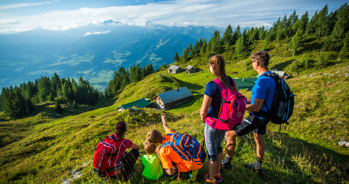 Eine Familie , bestehend aus zwei Söhnen, einer Tochter, Vater und Mutter, genießen die Außsicht vom Berg aufs Taal. Alle tragen Sportlichekleidung und bis auf den kleinsten Sohn tragen alle einen Wanderrucksack. Im Hintergrund befinden sich mehrer Almhütten und Bäume. In weiterer Entfernung erstreckt sich eine majestätische Bergfront.