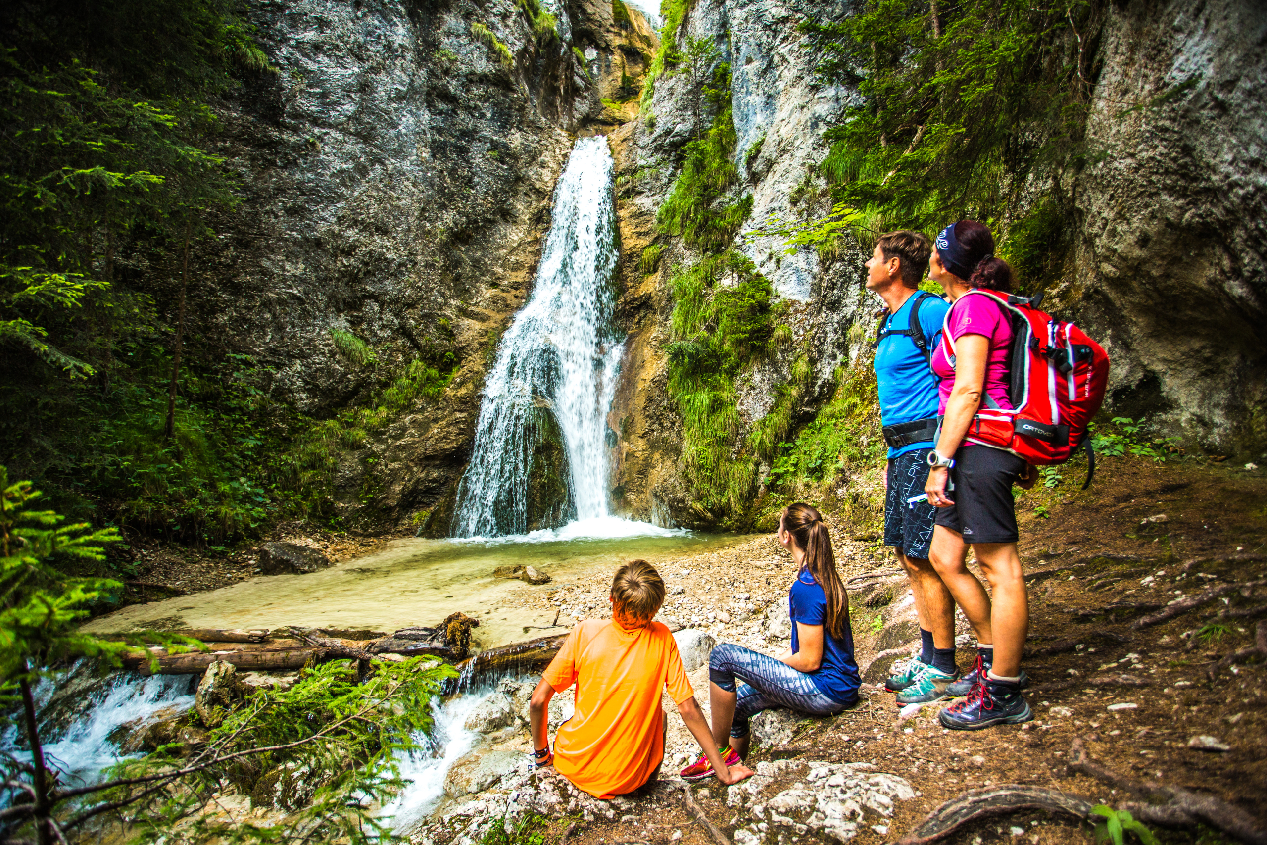 Eine Familie, bestehend aus Sohn und Tochter, sowie Mutter und Vater, machen einen Wanderausflug. Sie befinden sich gerade vor einem großem Wasserfall. Die Kinder sitzen und die Eltern stehen. Alle betrachten den hohen Wasserfall. Jeder von ihnen trägt spotliche Kleidung, whärend die Eltern jeweils einen Wanderrucksack tragen.