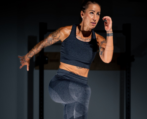 Bild zeigt eine Frau im mittleren Alter mit braunen Haaren und tätowieren Armen, im Sport-BH und Sportleggings, wie sie eine Übung vorzeigt.