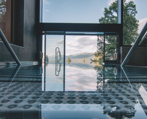 Ein luxoriöser, moderneer Innenbereichs des Infinity Pools mit spiegelndem Boden und großen Glasfronten, die einen weiten Blick auf den Pool, Wälder und Berge bieten.