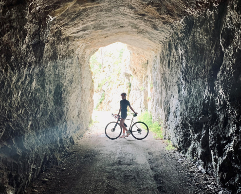 Das Bild zeigt eine Person mit einem Fahrrad, die in einem dunklen, felsigen Tunnel steht.