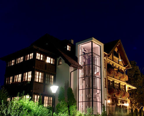 Das Hotel in der Nacht, mit erleuchteten Fenstern und einem modernen, gläsernen Außenlift in der Mitte, der sanft rosa leuchtet.