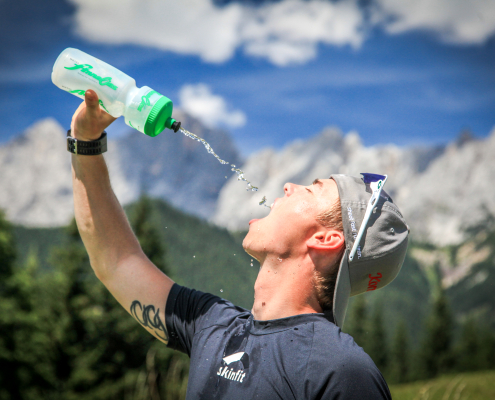 Ein junger,sportlicher Mann mit einer Kappe und darüber Sonnenbrille, trinkt Wasseraus einer Flasche, indem er sie über seinen weit geöffneten Mund hält.Das Wasser spritzt in einem Bogen. Er trägt Sportkleidung und hat eine Tätowierung am Arm. Im Hintergrund eine alpine Umgebung mit unscharfen Wäldern und Bergen.