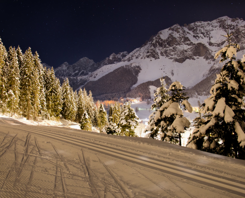 Eine Nachtaufnahme zeigt eine beleuchtete, präparierte Langlaufloipe, flankiert von schneebedeckten Tannenwäldern.Im Hintergrund erheben sich schneebedeckte Berge unter einem sternenklaren Nachthimmel. In der Ferne erkennt man die Lichter eines Dorfes.