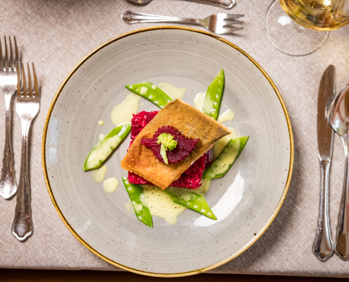 Ein Gourmet-Gericht liegt auf einem Teller: ein knuspriges Fischfilet auf rotem Püree, garniert mit Zuckerschoten und cremiger Soße.Der Teller ist umgeben von glänzendem Besteck auf einer hellen Tischdecke.