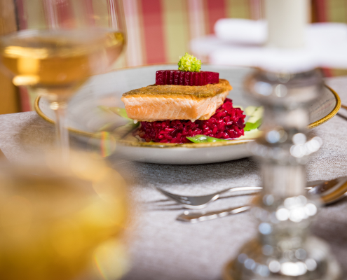Fokus auf ein Gourmet-Fischgericht (Lachs auf rotem Rote-Bete-Risotto) auf einem festlich gedeckten Tisch.