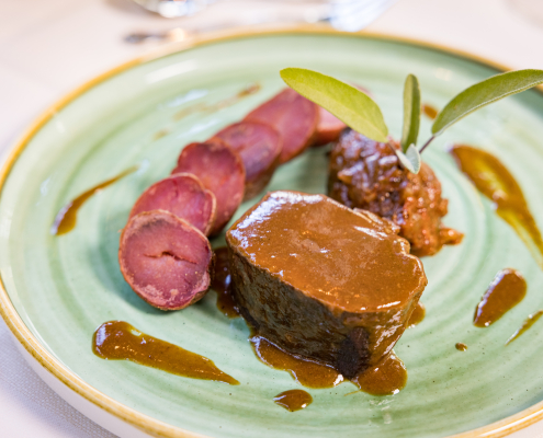 Nahaufnahme eines elegant angerichteten Gourmet-Hauptgangs auf einem hellgrünen Teller.Im Zentrum liegt ein geschmorrtes Stück Fleisch bedeckt mit einer glänzenden braunen Sauce. Daneben liegen Scheiben von lila Kartoffeln und dunkelbraune Ragout. Das Gericht ist mit Salbeiblättern garniert.