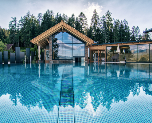 Infinity Pool des Hotel Annelies während eines Sonnenuntergangs. Im Hintergrund stehen Gebäude mit großen Glasfronten, die von Nadelbäumen umgeben sind.