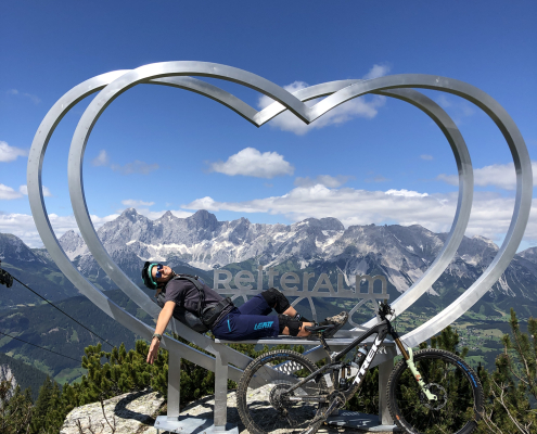 Mountainbikerin entspannt liegend auf einer Metallskulptur in Herzform , die auf einem Berggipfel platziert ist. Im Zentrum der Skulptur st der Schriftzug "ReiterAlm" zu erkennen. Im Hintergrund erstreckt sich eine majestätische Bergkette unter einem strahlend blauen Himmel mit einzelnen Wolken. Ihr Mountainbike ist an die Herzfigur angelehnt.
