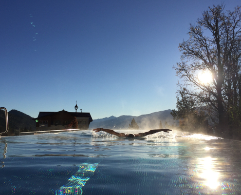 Silhouette eines Schwimmers beim Schmetterlingschwimmen in einem Infinity Pool, bei starkem Gegenlicht. Im Hintergrund ein traditionelles Haus, nebelverhangene Berge und ein kahler Baum bei Sonnenaufgang- oder untergang. Klarer blauer Himmel, luxuriöse, atmosphärische Szene.