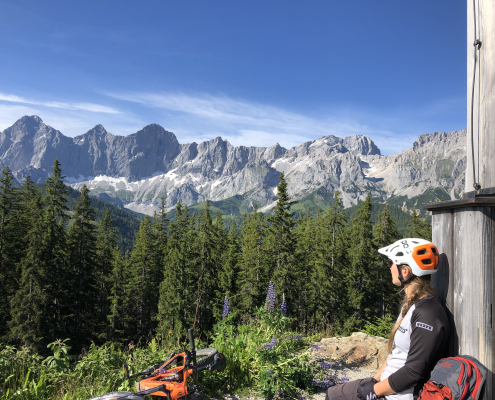 Eine Mountainbikerin sitzt und macht Pause, während sie den Anblick auf eine große Bergkette genießt.