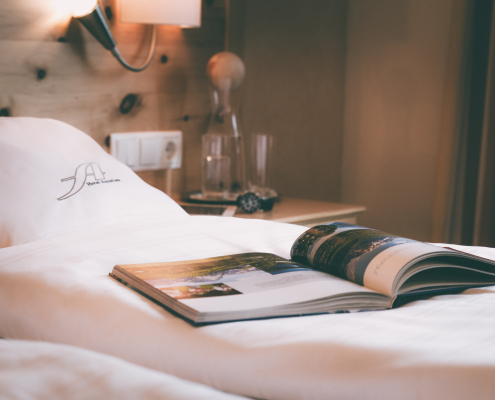 Ein aufgeschlagenes Magazin liegt auf einem weißen Holzbett vor einem Kissen. Im Hintergund ist ein Nachttisch aus Holz mit Gläsern und einer Lampe zu sehen , was eine gemütliche Atmosphäre schafft.