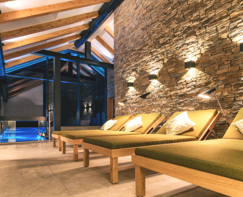 Ein luxuriöser Ruhebereich mit vier ölivgrünen Liegen vor einer beleuchteten Natursteinwand. Eine große Glasfront im Hintergrund gibt einen Blick auf einen blau beleuchteten Pool. Die Atmosphäre ist warm und entspannend.