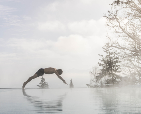 Eine Mann springt mit einem Kopfsprung in den beheizten Infinity Pool. Im Hintergrund stehen einige Bäume und der Himmel ist mit Wolen bedeckt.