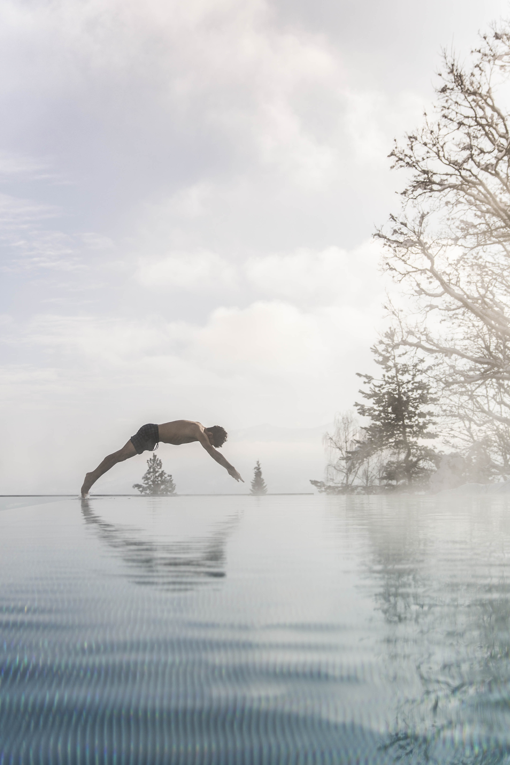 Schladming + Annelies Feb18-30 Eine Mann springt mit einem Kopfsprung in den beheizten Infinity Pool. Im Hintergrund stehen einige Bäume und der Himmel ist mit Wolen bedeckt.