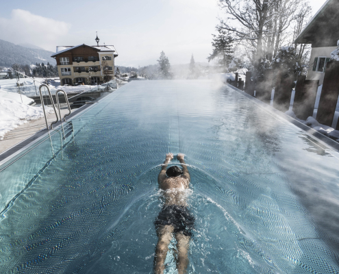 Eine Mann schwimmt mit gestreckten Körper Kraulbeine im Infinity Pool. Im Hintergrund zwei ältere Häuser und schneebedeckte Wälder.