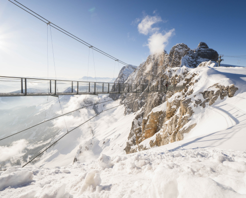 Eine spektakuläre Hängebrücke über einem Abgrund in den schneebedeckten Hochalpen, beleuchtet von der strahlenden Sonne unter blauem Himmel.