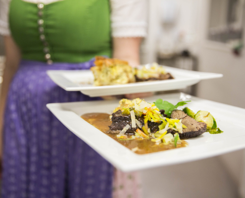 Eine Frau in einem Dirndl serviert zwei festlich angerichtete Hauptspeisen auf weißen, rechteckigen Tellern Auf beiden Tellern ist jeweils 2 Stückefleisch mit einem Saft und feingeschnittenes Gemüse.