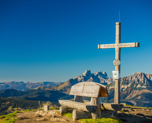 Malerisches Bergpanorama unter einem klaren,tiefblauen Himmel. Im Vordergrund stehen ein hölzernes Gipfelkreuz und eine rustikale Holzbank.Auf dem Querbalken des Kreuzes steht eine Höhenangabe (2001m) und der Name des Gipfels ( Gassel Höh). Im Hintergrund erstreckt sich eine atemberaubende Berglandschaft.
