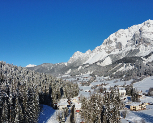 Panoramablick auf ein verschneites Alpendorf in einem Tal, eingerahmt von dichtem Nadelwald und einem riesigen, schneebedeckten Berggipfel unter blauem Himmel.