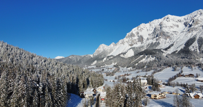 Winterlandschaft Panoramablick auf ein verschneites Alpendorf in einem Tal, eingerahmt von dichtem Nadelwald und einem riesigen, schneebedeckten Berggipfel unter blauem Himmel.