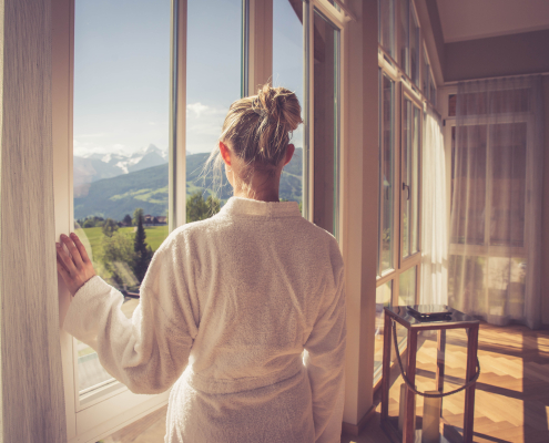 Eine Frau von hinten, in einem weißen Bademantel, steht vor einem großen Fenster und blickt auf eine atemberaubende Alpenlandschaft mit grünen Tälern und schneebedeckten Berggipfeln. Das Sonnenlicht fällt durch das Fenster in den hellen Raum.