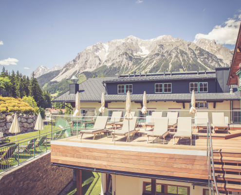 Sonnige Holzterrasse des Hotels, ausgestattet mit Liegenstühlen, Sonnenschirmen und einem atemberaubenden Ausblick auf eine Bergkette.