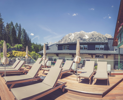 Sonnige Holzterrasse des Hotels, ausgestattet mit Liegenstühlen, Sonnenschirmen und einem atemberaubenden Ausblick auf eine Bergkette.