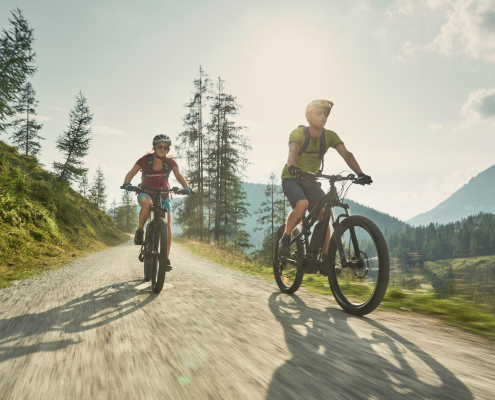 Zwei Mountainbiker (ein Mann und eine Frau) fahren auf einem sonnigen Schotterweg bergab in einer alpinen Berglandschaft. Die Sonne strahlt hell über den Baumwipfeln, und im Hintergrund sind grüne Hänge und Berge zu sehen. Die Biker tragen Helme, Rucksäcke und sportliche Kleidung.