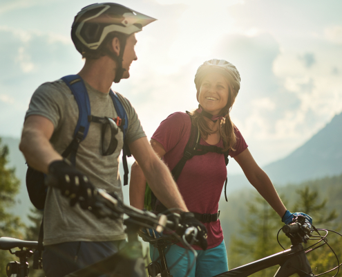 Glückliches Mountain-Biker Paar mit Helmen,das sich gegenseitig anlächelt. Sie schieben beide ihrer Räder nebeneinander her. Im Hintergund eine unscharfe Natur - Wälder und Berge
