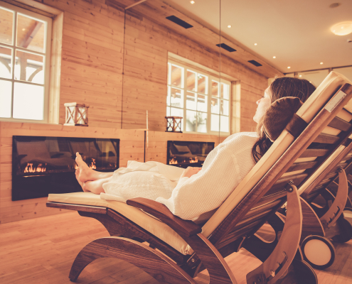 Eine Frau in einem Bademantel liegt entspannt auf einer Holzliege in einem Ruheraum. Der Raum ist holzgetäfelt und verfügt über zwei integrierte Kamine und großer Fenster, was eine warme und ruhige Wellness-Atmosphäre schafft.