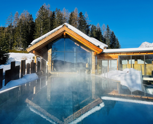 Ein dampfender Infinity-Außenpool vor einem modernen Holz-Glas-Gebäude in einer schneebedeckten alpinen Umgebung unter blauem Himmel.