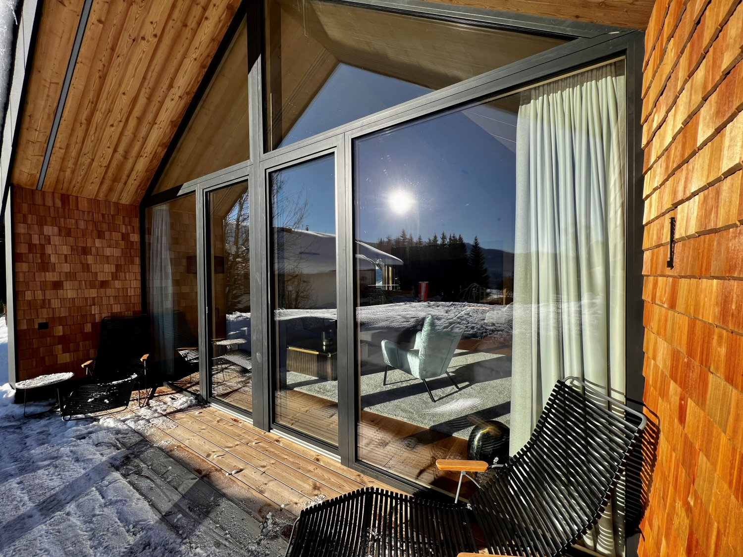 Eine moderne Holzwand mit großer Glasfront bietet den Blick von einer kleinen, verschneiten Terrasse in einen Raum mit modernen Sitzgelegenheiten.