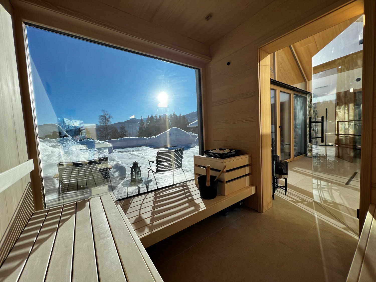 Blick aus einer hellen Holzsauna durch eine große Fensterfront auf eine schneebedeckte Terrasse bei Sonnenschein. Rechts ist der Zugang zu einem angrenzenden Wellnessbereich.