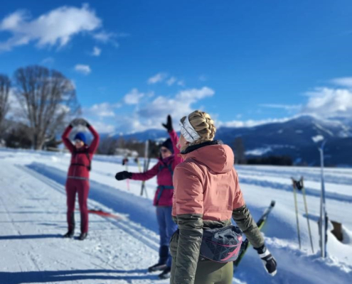 Eine Langlauf-Trainerin macht mit 2 Auszubildenden eine Aufwärmübung. Sie stehen auf einer Loipe unter blauem Himmel.