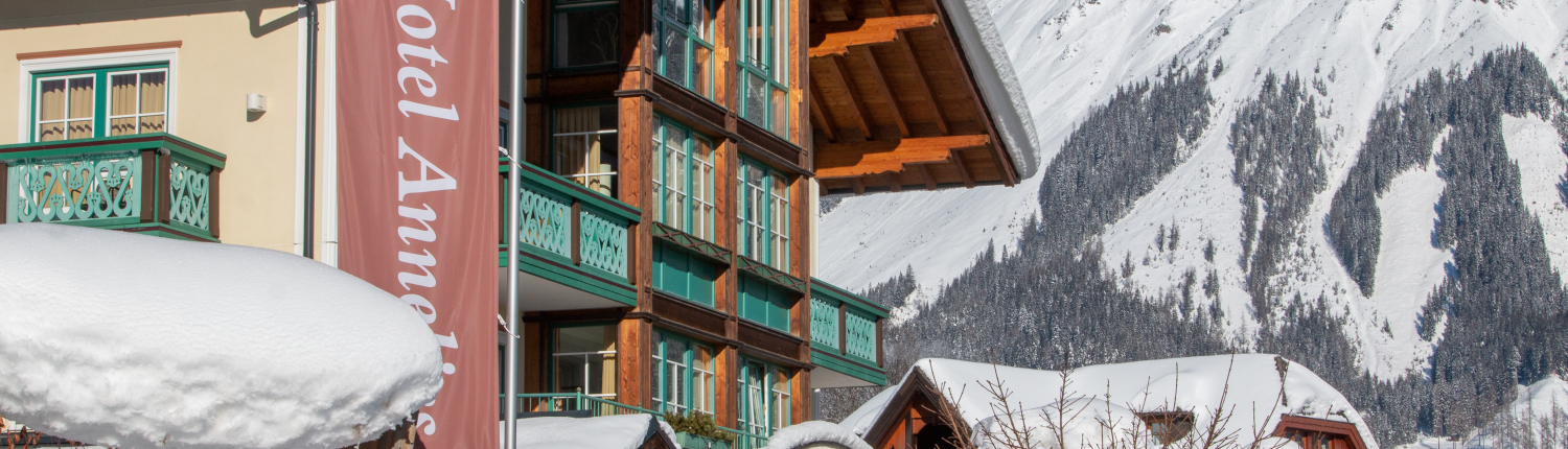 Die Fassade des Hotel Annelies ist in einer tief verschneiten Winterlandschaft zu sehen. Im Vordergrund steht ein Hotel-Banner, und direkt im Hintergrund erheben sich hohe, schneebedeckte Berge.