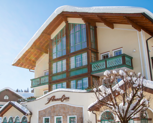 Das Bild zeigt die Hauptfassade des Hotel Annelies an einem sonnigen Wintertag, mit einer großen Glasfront, grünen Balkonen und Schnee auf dem Dach.