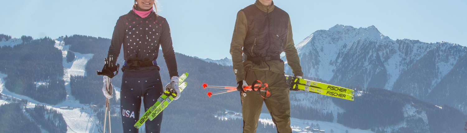 Zwei Langläufer (Mann und Frau) in Skibekleidung, die bei Sonnenschein auf einer verschneiten Loipe in einer alpinen Winterlandschaft gehen.