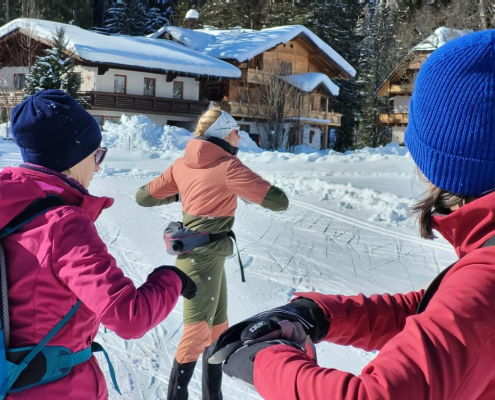 Eine Langlauflehrerin im olivgrün-rosa Anzug demonstriert eine Übung für zwei Schüler (in pinker und roter Jacke) auf einer verschneiten Loipe vor Alpenhäusern.