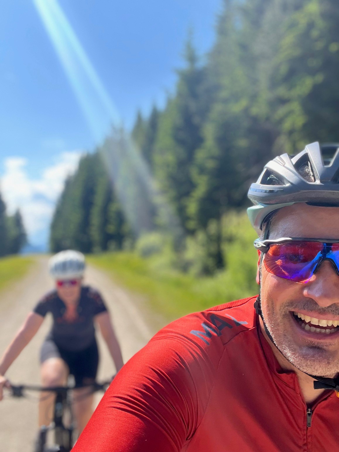 Vorne ist ein Mann in rotem Trikot, mit Helm und Sonnenbrille, der lächelnd in die Kamera schaut. Hinter ihm fährt eine Frau in dunkler Sportkleidung und Helm, leicht verschwommen im Hintegrund. Um sie herum sind grüne Bäume und blauer Himmel mit Sonnenschein.