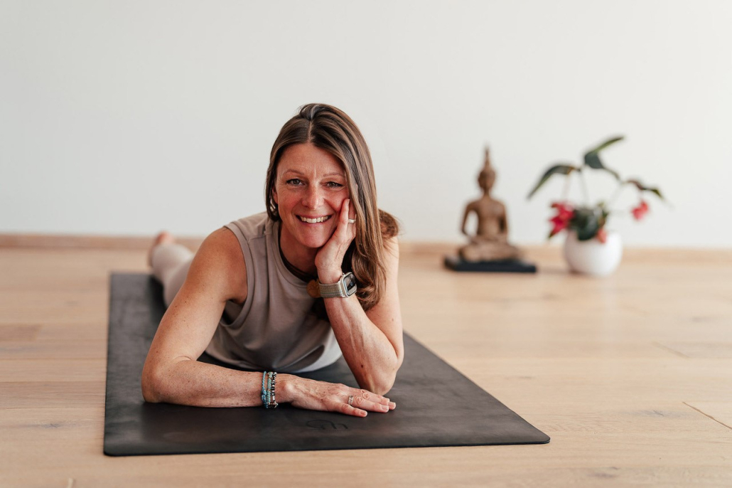 Eine Frau im mittleren Alter liegt mit normaler Altagskleidung und offenen Haaren auf einer Jogamatte. Im Hintergund befinden sich eine kleine Buddha-Statue und ein Blumentopf mit Blumen.
