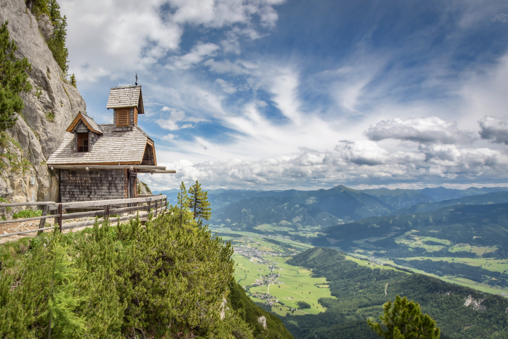 Kapelle mit Top Aussicht Das Bild zeigt eine kleine, rustikale Holzkapelle an einem steilen Berghang. Von dort aus hat man einen weiten Blick über ein Tal mit DörfernWäldern und dahinter Berge.
