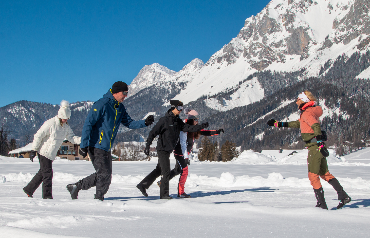 Langlaufschule: Training Ein Profi bringt vier Personen in Winterkleidung das Langlaufen ohne Skier auf einer Langlaufloipe bei. ImHintergund sind schneebedeckte Berge zu sehen.