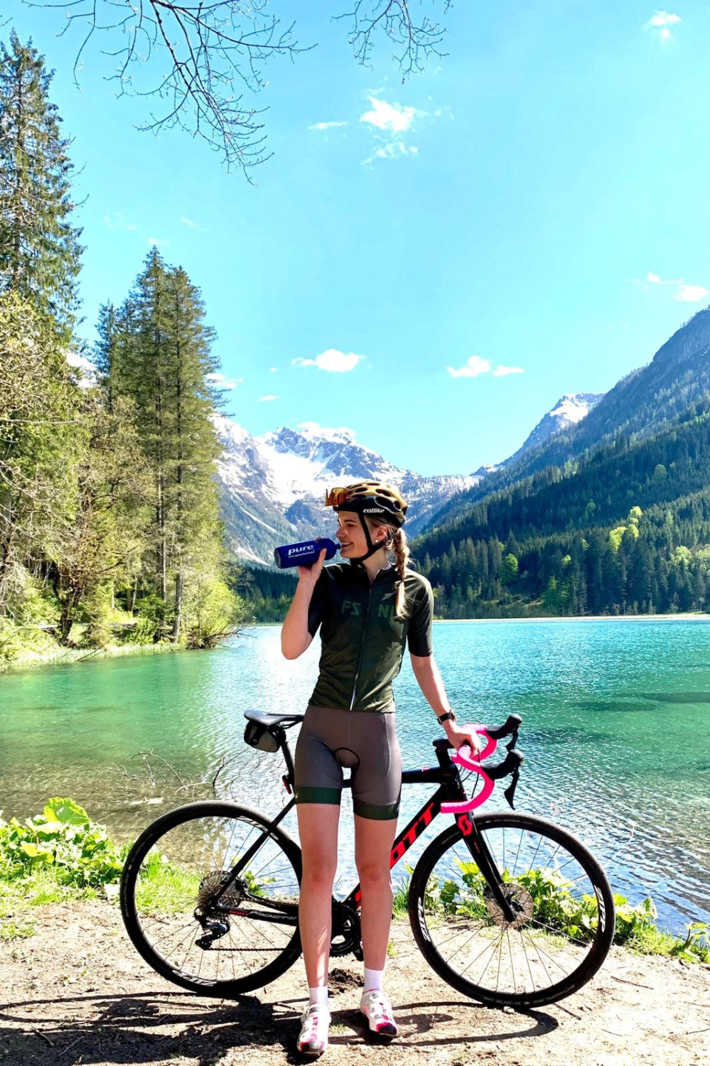 Das Bild zeigt eine junge Frau in Radsportbekleidung, die eine Pause an einem See in den Bergen macht. Sie steht neben ihrem Rennrad und trinkt aus einer blauen Trinkflasche. Sie trägt einen Helm und hat die Haare zu einem Zopf geflochten.
