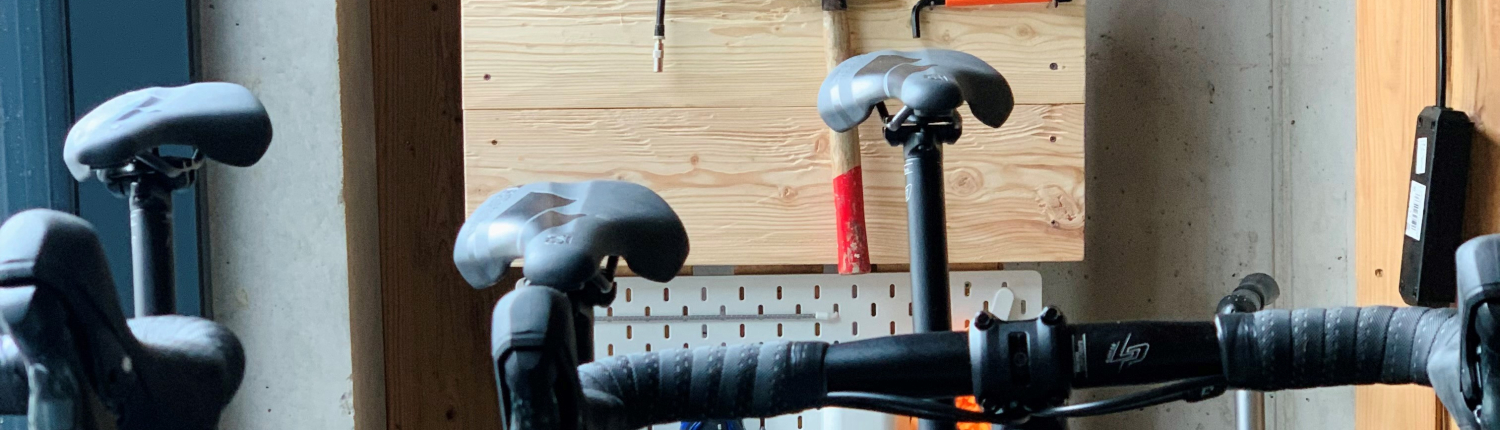 Das Bild zeigt einen Innenraum, der als Fahrrad-Werkstatt genutzt wird. Im Vordergrund sind Lenker und Sättel von mehreren Fahrrädern zu sehen. An der Rückwand hängt auf einer hellen Holztafel eine Vielzahl von Werkzeugen für die Fahrradreparatur, darunter Zangen, ein Hammer und eine Luftpumpe.